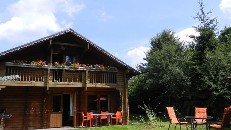 Ein gemütliches Holzhaus mit einem Balkon voller Blumen. Im Vordergrund stehen orangefarbene Stühle und ein Tisch auf der grünen Wiese.