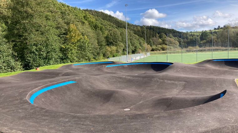 Eine moderne Bike-Piste mit hügeligem Terrain und blauen Markierungen. Im Hintergrund sind grüne Bäume und ein Sportfeld zu sehen.
