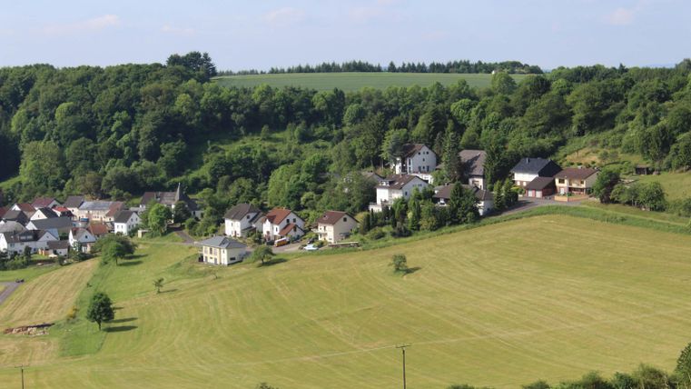 Eine malerische Landschaft mit einem kleinen Dorf und grünen Wiesen. Umgeben von sanften Hügeln und Bäumen.