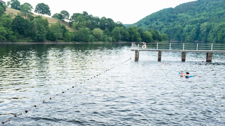 Ein ruhiger See mit grünen Hügeln im Hintergrund. Auf der Wasseroberfläche schwimmt eine Person in der Nähe eines Holzstegs.