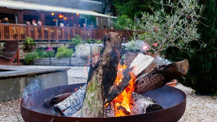 Ein gemütliches Lagerfeuer brennt in einem großen Metallfeuerkorb. Im Hintergrund sind eine Terrasse und Pflanzen zu sehen, die eine einladende Atmosphäre schaffen.