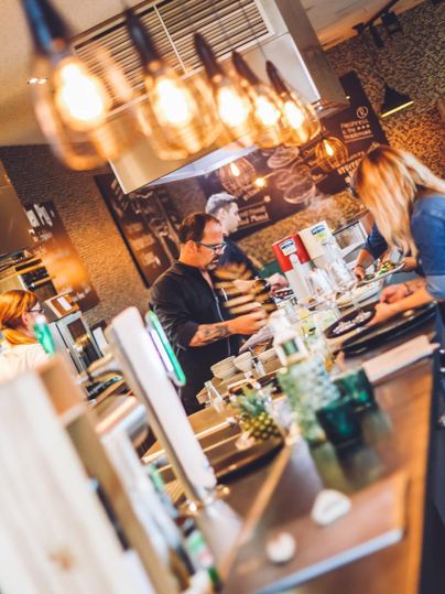 Eine belebte Café-Szene mit Baristas, die Getränke zubereiten. Gemütliche Atmosphäre mit warmen Lichtern und zahlreichen Pflanzen.