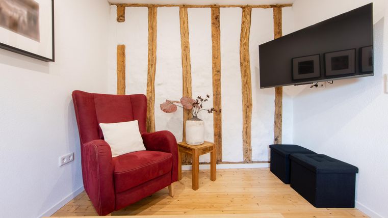 Ein gemütlicher Wohnraum mit einem roten Sessel und einem kleinen Holztisch. An der Wand sind sichtbare Holzstrukturen und ein Fernseher angebracht.