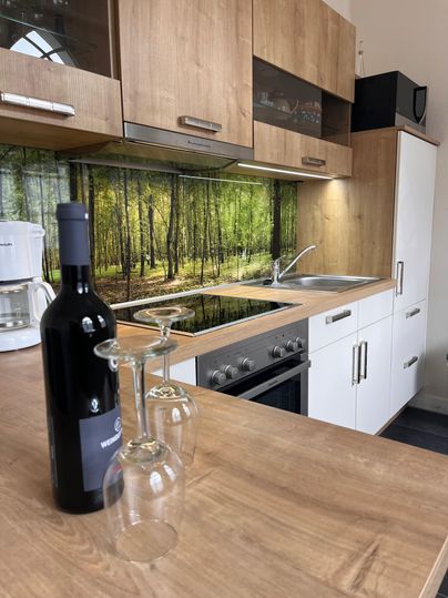 Eine moderne Küche mit Holzschränken und einem ansprechenden Hintergrundbild von einem Wald. Auf der Arbeitsplatte stehen eine Flasche Wein und Weingläser bereit.