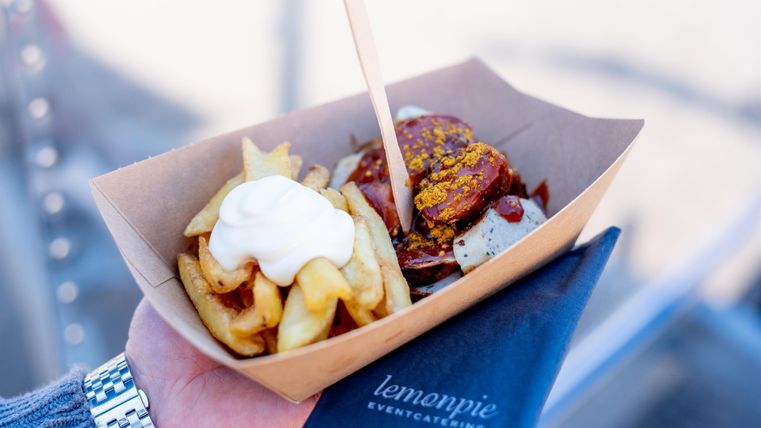 Een handgerecht met knapperige frietjes en een portie saus. De gerechten zitten in een papieren verpakking en worden door iemand vastgehouden.