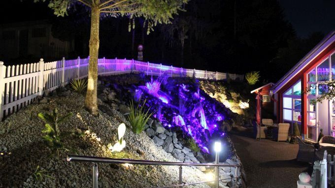 Ein schöner Gartenweg mit bunten Lichtern und einem Wasserfall. Die Umgebung ist nachts beleuchtet und wirkt einladend.