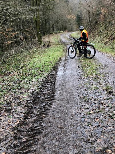 A mountain biker stands on a muddy forest path. The surroundings are surrounded by trees and depict a rural atmosphere.