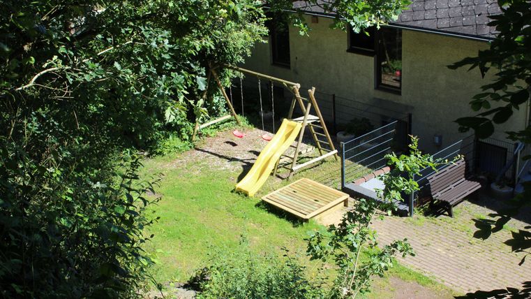 Ein Spielplatz im Garten mit einer Rutsche und einer Schaukel. Umgeben von Bäumen und einem hellen, sonnigen Himmel.