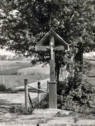Een houten wegkruis staat langs een landweg, omringd door bomen en weiden. Op de achtergrond strekt zich een open landschap uit.