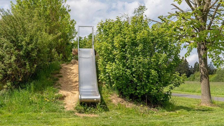 Een glijbaan van metaal staat in een groen park. Omringd door struiken en bomen, nodigt ze uit om te spelen.