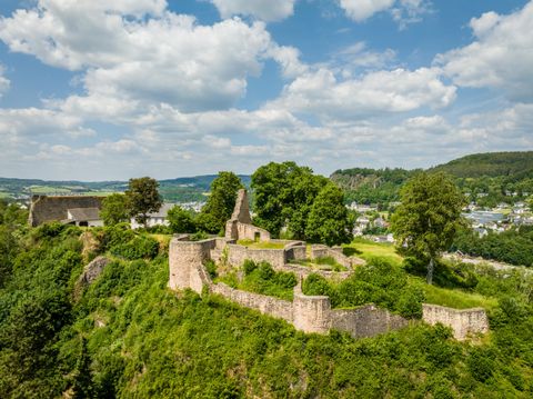 Luftaufnahme der Löwenburg in Gerolstein, umgeben von grüner Landschaft und Bäumen. Im Hintergrund ist die Stadt zu sehen, unter einem blauen Himmel mit Wolken.
