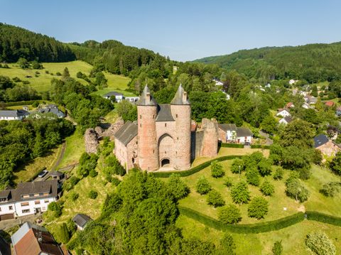 Luftaufnahme der Bertradaburg in Mürlenbach, umgeben von grüner Landschaft und Häusern. Die Burg hat zwei markante Türme.