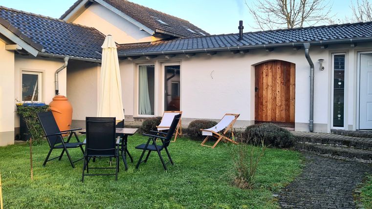 Ein gemütlicher Garten mit Gartenstühlen und einem Sonnenschirm. Im Hintergrund ist ein helles Haus mit Holzdetails zu sehen.