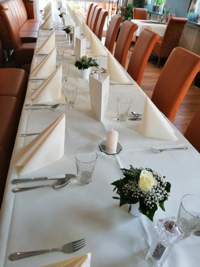 Ein festlich gedeckter Tisch mit weißen Tischtüchern, Servietten und Blumenarrangements. Die Stühle sind in einem warmen Braunton gehalten und schaffen eine einladende Atmosphäre.