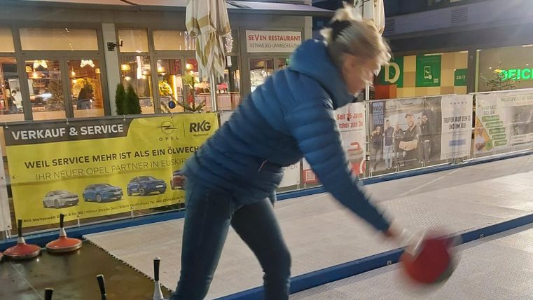 Eine Frau spielt im Freien Bowling an einer beleuchteten Bahn. Hintergrund mit dem Wort "SEVEN" und Werbeplakaten.