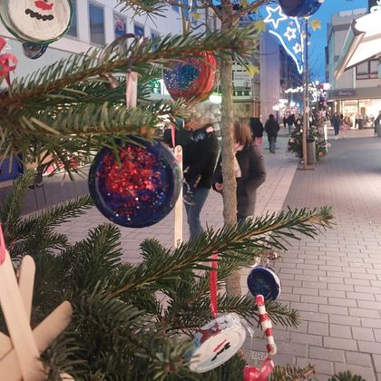 Ein geschmückter Weihnachtsbaum mit bunten Ornamenten. Im Hintergrund sind Menschen auf einer festlich beleuchteten Straße zu sehen.