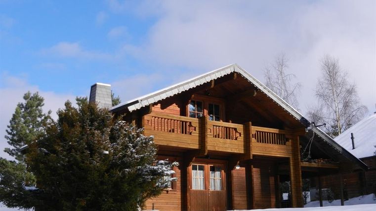 Ein gemütliches Holzhaus im Schnee mit einem schönen Balkon. Umgeben von schneebedeckten Bäumen und einem blauen Himmel.