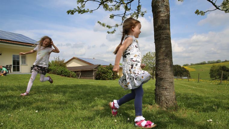 Twee kinderen spelen vrolijk in het gras onder een boom. Op de achtergrond zijn een huis en een groene landschap te zien.