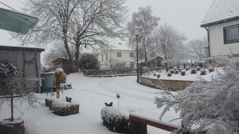 A snow-covered landscape with houses in the background. The road and the garden are completely covered in snow.