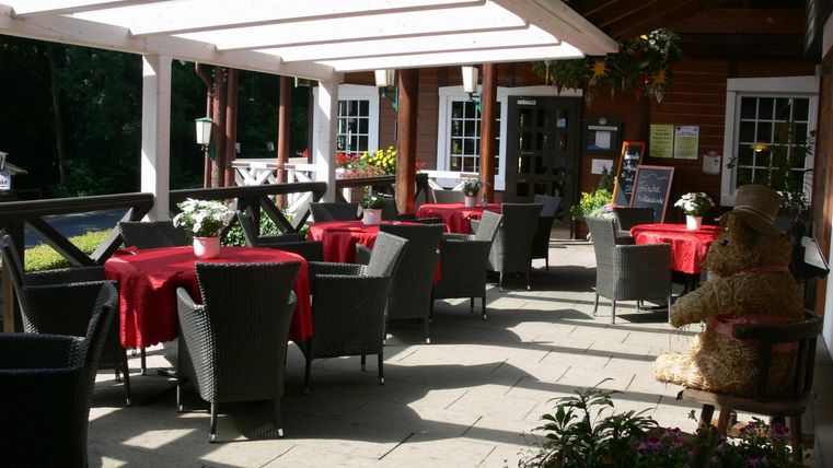 Eine gemütliche Terrasse mit Tischen und Stühlen, die mit roten Tischdecken dekoriert sind. Im Hintergrund steht eine dekorative Figur aus Stroh.