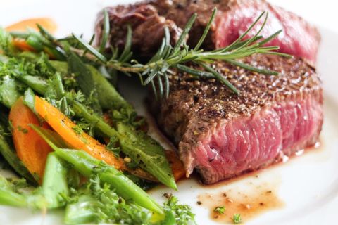 A juicy piece of beef steak with spices and fresh rosemary, served on a plate. Next to it are colorful vegetables like green beans and carrots.