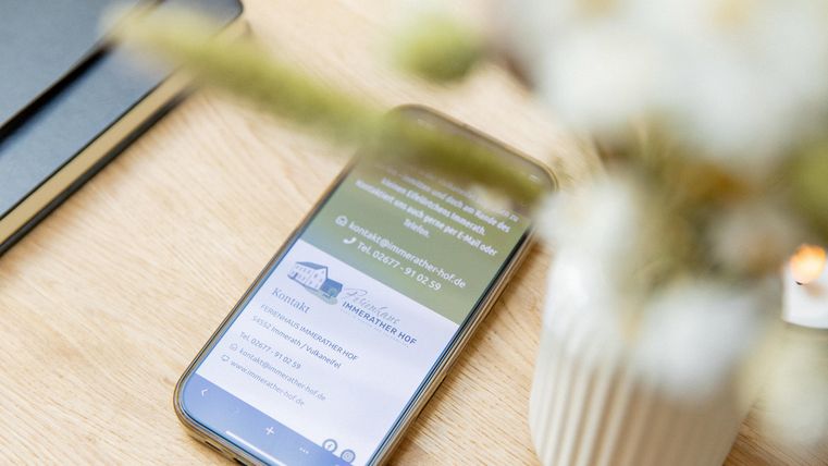 Ein Smartphone liegt auf einem Tisch, umgeben von einem Notizbuch und einer Blumenvase. Die Anzeige zeigt eine Website mit Informationen.