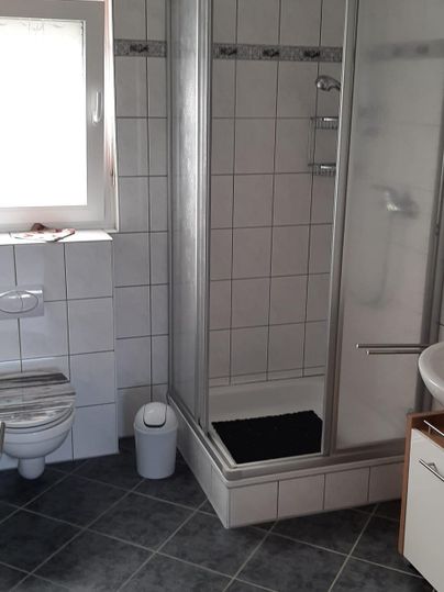 Ein Badezimmer mit einer Dusche, Toilette und einem Waschbecken. Die Wände sind gefliest und es gibt ein Fenster für Tageslicht.