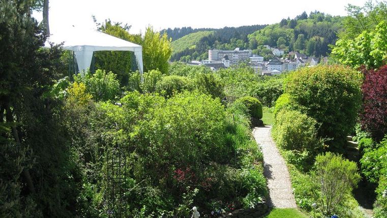 Een mooie tuin met weelderig groen en kleurrijke planten. Op de achtergrond is een pittoresk landschap met huizen en heuvelachtige bossen te zien.