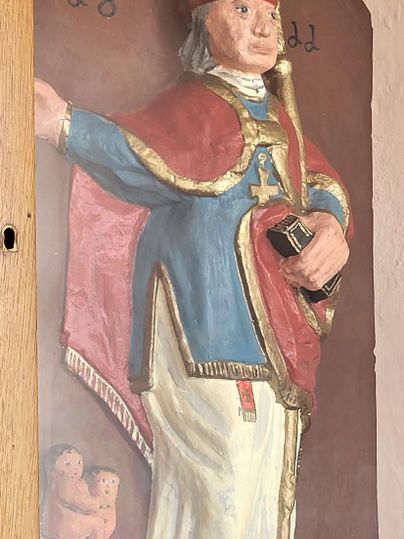 Eine bemalte Statue von St. Nikolaus in einem festlichen Gewand. Er hält ein Buch und steht vor einer dekorativen Rückwand.