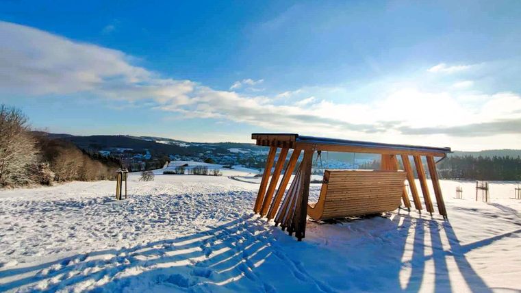 Eine ruhige Winterlandschaft mit viel Schnee und einem Holzschaukelstuhl im Vordergrund. Die Sonne scheint und schafft eine warme Atmosphäre.
