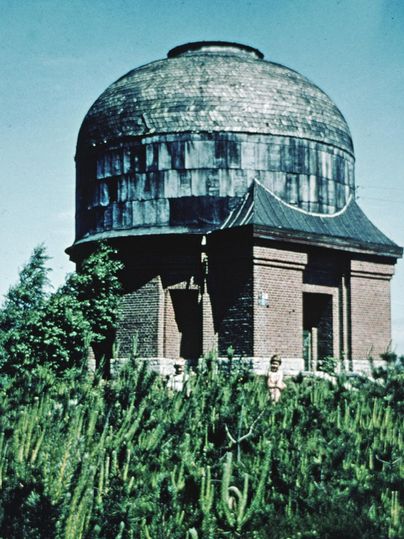 A historical observatory with a domed roof stands before a green landscape. Various plants grow around the building.