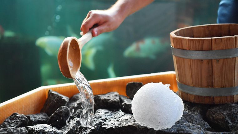 Een houten lepel giet water over donkere stenen in eencontainer. Naast de stenen ligt een ronde ijssculptuur.