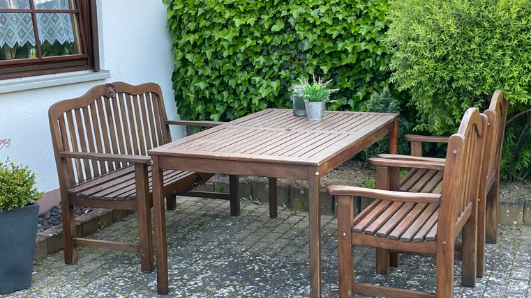 Een houten dienblad met vier stoelen staat buiten. Op de achtergrond zijn groene struiken te zien.