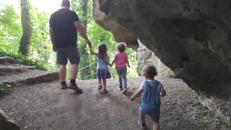 Eine Person geht mit zwei kleinen Kindern auf einem Weg in einem Wald. Sie halten sich an den Händen und gehen in Richtung Felsformationen.