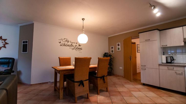 A bright dining room with a large table and chairs. The walls are painted in neutral colors, and the kitchen adjoins the living area.