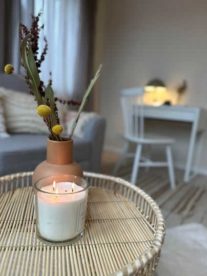 Een gezellige woonomgeving met een kaars en een vaas op een tafel. Op de achtergrond is een lichte kamer zichtbaar met een bureau en een stoel.