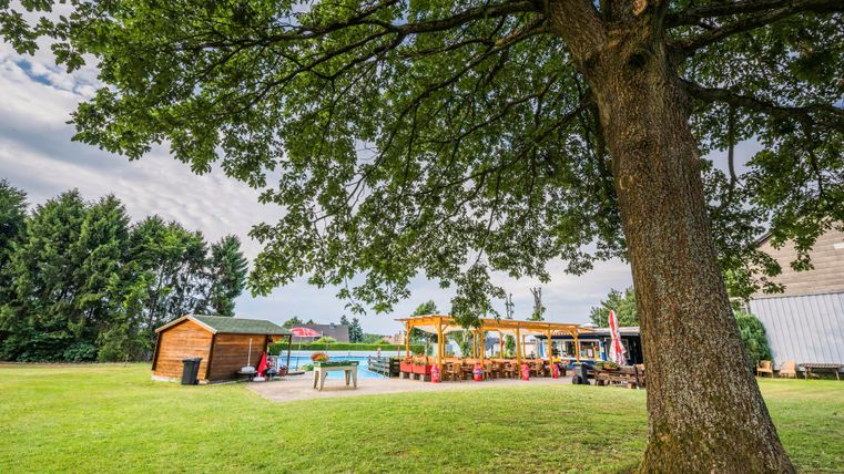 Eine grüne Wiese mit einem großen Baum und einer Holzlaube. Im Hintergrund ist ein Restaurantbereich mit Sitzgelegenheiten zu sehen.