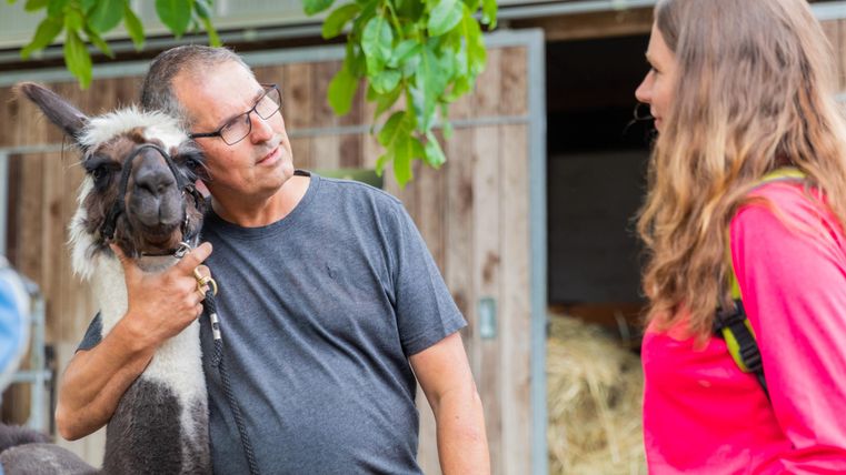 Ein Mann spricht mit einer Frau, während er ein Lama streichelt. Beide lächeln und scheinen sich zu unterhalten.