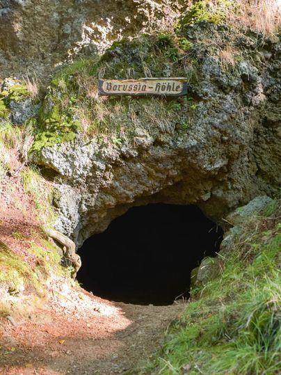 Entrance to the Borussia Cave, surrounded by moss-covered rocks and grass. A wooden sign with the inscription "Borussia Cave" is visible.