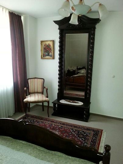 Ein Schlafzimmer mit einem großen, dekorativen Spiegel und einem eleganten Stuhl. Die Wände sind hell, und ein Teppich verleiht dem Raum ein gemütliches Ambiente.