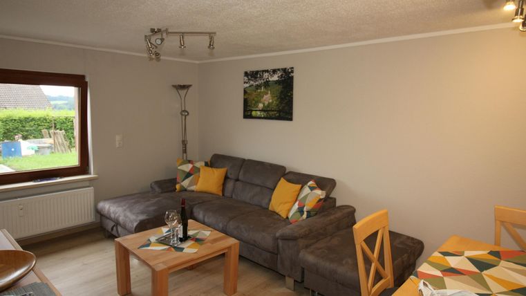 Een gezellige woonkamer met een grijze bank en kleurrijke kussens. De ruimte is helder en uitnodigend, met een tafel en een plant.