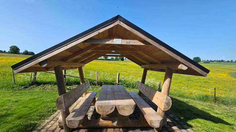 Ein Holzunterstand mit einem großen Tisch und Bänken steht auf einer Wiese. Im Hintergrund sind grüne Felder und ein klarer blauer Himmel zu sehen.