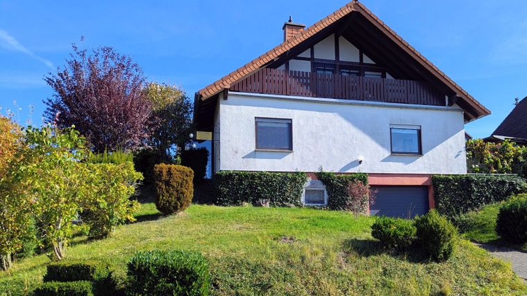 Een mooi huis op een heuvel met een verzorgde tuin. De zon schijnt en de lucht is blauw.