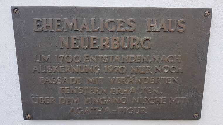 Eine Metalltafel mit der Aufschrift „Ehemaliges Haus Neuerburg“. Sie liefert Informationen über das Gebäude, das um 1700 entstand und 1970 nur noch als Fassade existiert.