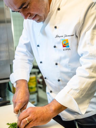 Ein Koch, der frische Kräuter auf einem Schneidebrett choppt. Er trägt eine weiße Kochjacke und arbeitet in einer modernen Küche.