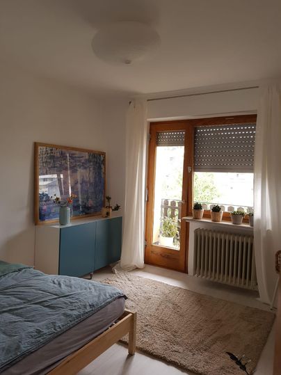 Een lichte slaapkamer met een bed, een bureau en grote ramen. Op de vensterbank staan planten en de ruimte voelt vriendelijk en uitnodigend aan.