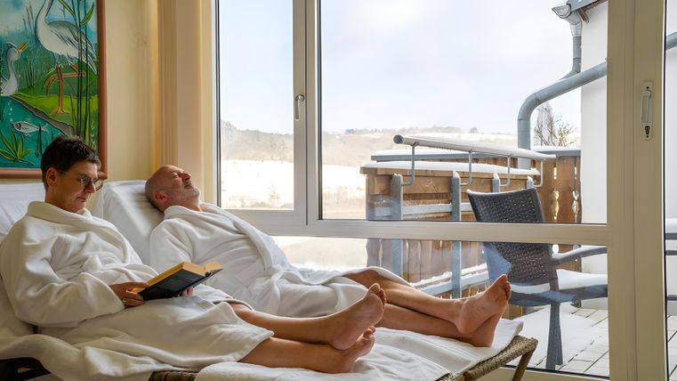 Zwei Personen in Bademänteln entspannen auf Liegen. Durch das Fenster ist ein ruhiger Ausblick auf eine Terrasse zu sehen.