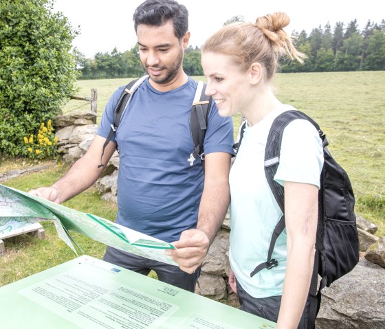 Twee mensen met rugzakken bekijken een wandelkaart voor een groene weide en bomen., © Eifel Tourismus GmbH, AR-shapefruit AG
