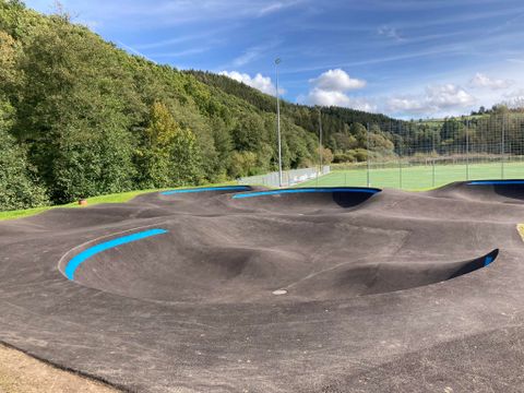 Eine moderne Bike-Piste mit hügeligem Terrain und blauen Markierungen. Im Hintergrund sind grüne Bäume und ein Sportfeld zu sehen.