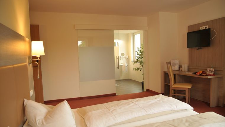 Ein modernes Hotelzimmer mit zwei Einzelbetten, einem Schreibtisch und einem Fernseher. Badezimmer und Fenster sorgen für viel Tageslicht.